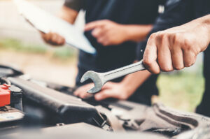 A mechanic is holding a wrench and working on a car. Another person in the background is holding a clipboard