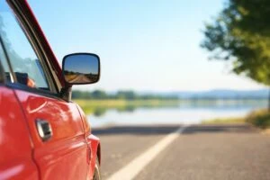 Close-up of a red car's side mirror and door on a rural road with a lake and trees in the background.